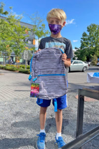 A boy wears a face mask, stands in front of a blue sky, and holds up a gray striped backpack