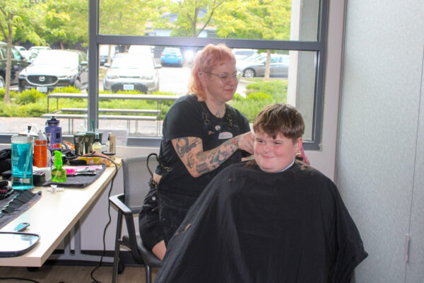 A stylist with pink hair cuts the hair of a boy at the Cascadia school drive.