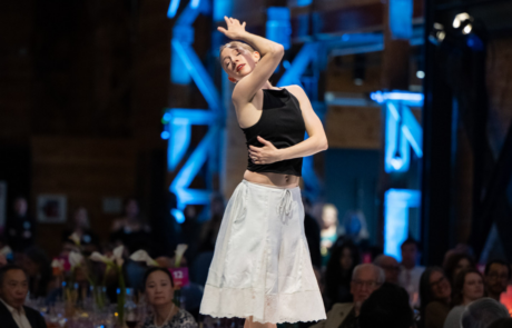 Female dancer wearing a black sleeveless top and white skirt performs on a blue-lit stage with an audience watching in the foreground.