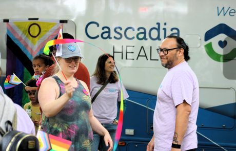 Group of people at a Pride event holding rainbow flags in front of a vehicle with Cascadia Health branding on it.