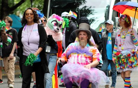 Group of people at a Pride parade wearing colorful outfits and accessories, smiling and walking together down a street.