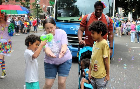 Children and adults at a street parade blowing bubbles beside a parked blue bus on a sunny day.