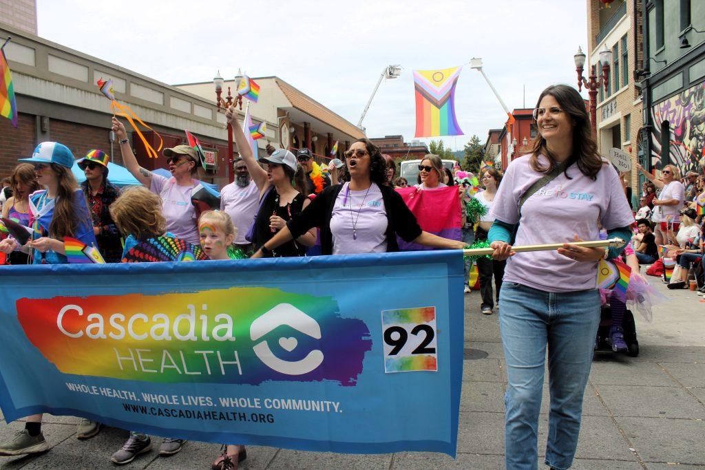 People marching in the Portland Pride parade holding rainbow flags, with a Cascadia Health banner in front. It says, Whole Health. Whole Lives. Whole Community.