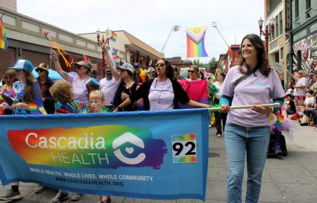 People marching in the Portland Pride parade holding rainbow flags, with a Cascadia Health banner in front. It says, Whole Health. Whole Lives. Whole Community.