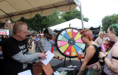 People gather at Cascadia Health's booth, a person with tattoos and pink hair listens to a staff member who is operating a colorful wheel used for trivia questions.