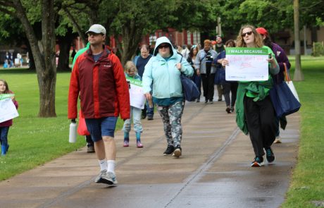 People walking in a park during a charity walk, holding signs that read 'I walk because nobody should walk alone' and other supportive messages.