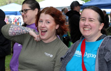 Two Cascadia Health staff posing and smiling at an outdoor event, with tents in the background and green surroundings.