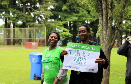 Two smiling walkers in a park, one holding a green sign that reads 'I WALK BECAUSE... THERE IS A NEED' with trees in the background