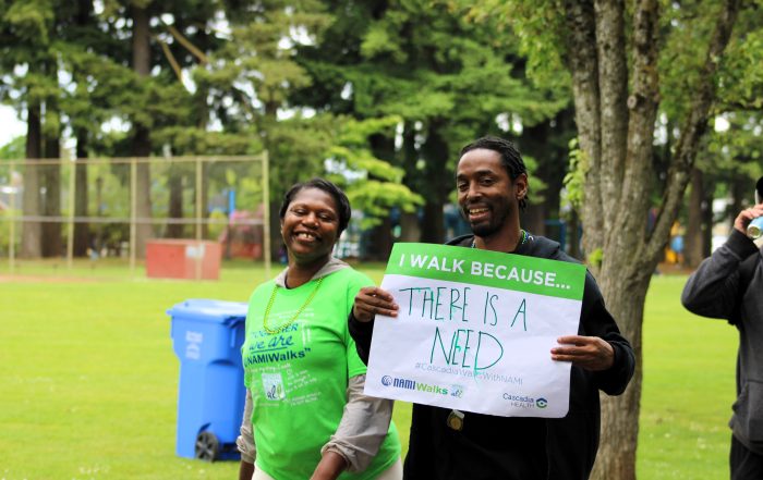 Two smiling walkers in a park, one holding a green sign that reads 'I WALK BECAUSE... THERE IS A NEED' with trees in the background