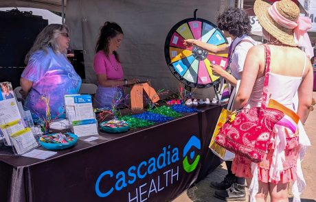 Health fair booth with a large colorful prize wheel, volunteers at a table handing out brochures and prizes labeled Cascadia Health.