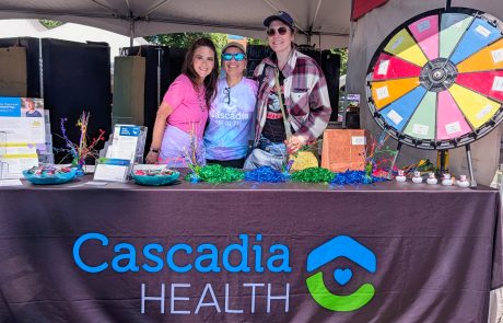 Three smiling people stand behind a Cascadia Health booth at a health fair, with brochures, colorful decorations, and a large spinning prize wheel to the side.