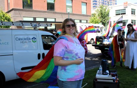 Person wearing a colorful tie-dye shirt and rainbow cape at a Pride parade, smiling with flags in the background near a Cascadia Health van.