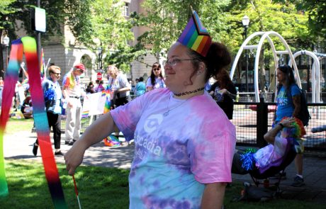 Person wearing a rainbow party hat and tie-dye shirt at a Pride parade outdoors, smiling with colorful streamers nearby.