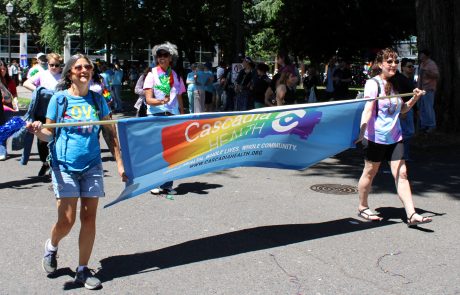 Group of parade participants carries a long blue rainbow banner with 'Cascadia Health' text, smiling faces in sunny outdoor crowd.