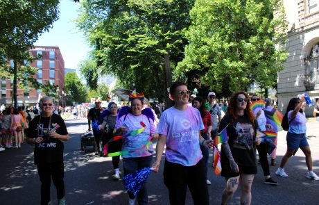 People marching in a Pride parade along a sunny city street with rainbow flags and colorful outfits under leafy trees.