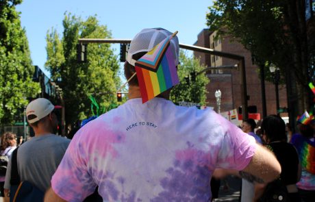 Back view of a Pride parade participant wearing a rainbow flag hat and pink-purple tie-dye shirt, with 'HERE TO STAY' on the back, in a city street.