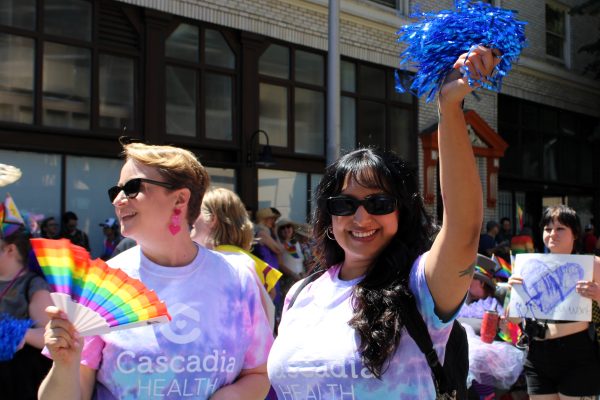 Group of parade participants waving rainbow flags and wearing tie-dye shirts during a city Pride march, smiling at the camera.