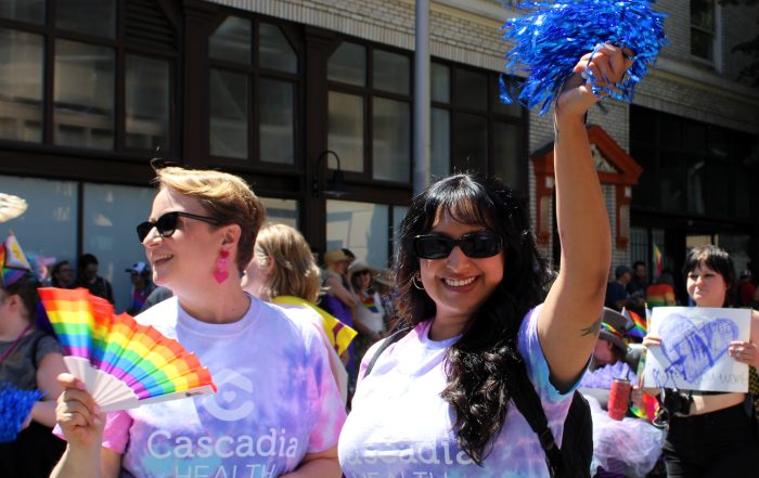 Group of parade participants waving rainbow flags and wearing tie-dye shirts during a city Pride march, smiling at the camera.