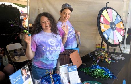 Two women in tie-die Cascadia Health shirts wave, standing behind a booth with print collateral, safety supplies, and a large carnival-style trivia wheel.
